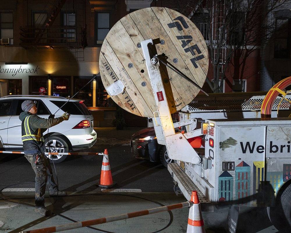 A worker guides new fiber-optic cable from a reel truck into a city street at night